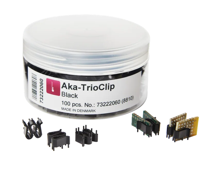 Akasel Aka-TrioClip Black i en transparent plastbehållare med vitt lock. Etiketten lyder “Aka-TrioClip Black,” “100 pcs. No.: 73222060 (8810),” och “MADE IN DENMARK.” Svarta plastclips och inbäddade prover med gröna och gula kretskort visas i förgrunden, speciellt utvecklade för metallografiska applikationer.